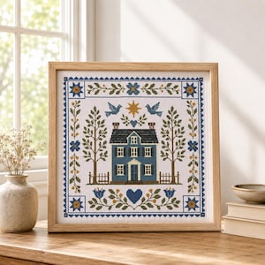 May include: A square cross-stitch embroidery in a light wood frame featuring a blue house with a dark roof, flanked by two tall trees. The design includes blue birds, a yellow sun, floral borders, and a blue heart, all on a white fabric background.