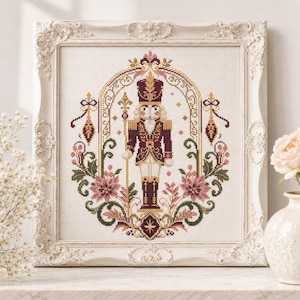 May include: A cross-stitch embroidery of a nutcracker soldier in a maroon and gold uniform, holding a staff. The figure is framed by an ornate archway with floral accents in shades of pink, green, and gold, all encased in a white vintage frame.