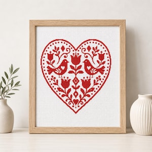 Op de afbeelding: Een ingelijst kruissteekkunstwerk met een rood hartontwerp met vogels, bloemen en bladeren. Het kunstwerk is geplaatst tegen een witte achtergrond en ingelijst in een licht houten frame. Een decoratief item.