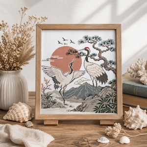 May include: A square cross-stitch embroidery in a light wood frame depicting two red-crowned cranes standing near a pine tree and mountains against a large orange sun. The scene includes small flying birds and stylized clouds in a detailed needlework design.