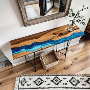 Puede incluir: Una consola de madera con diseño de río. La mesa presenta una tapa de madera natural con un río de resina epoxi azul. Tiene patas de metal negro y una cesta tejida debajo. Un espejo y un jarrón con vegetación están en el fondo.