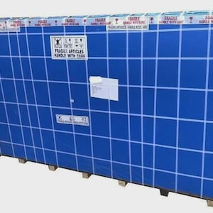 May include: Large, rectangular package wrapped in blue plastic with a grid pattern. The package is labeled "FRAGILE ARTICLES HANDLE WITH CARE" with warning symbols and tape. It is secured to a wooden pallet.