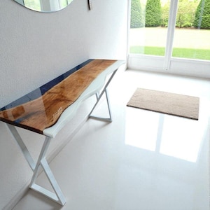 May include: A modern console table with a live edge wooden top, featuring a glossy finish and a blue resin inlay. The table has a white, wavy edge and is supported by white, X-shaped metal legs. The table is approximately 152 cm long.