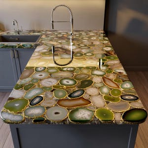 May include: A kitchen countertop made of agate stone slices in shades of green, white, and brown, with gold-colored edging. The countertop features a stainless steel faucet and a sink. The cabinet is painted in a gray color.