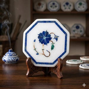 May include: White marble coaster with a blue floral inlay, set on a wooden stand. The coaster has an octagonal shape with a blue border. The floral design features a blue flower and green leaves. Decorative item.