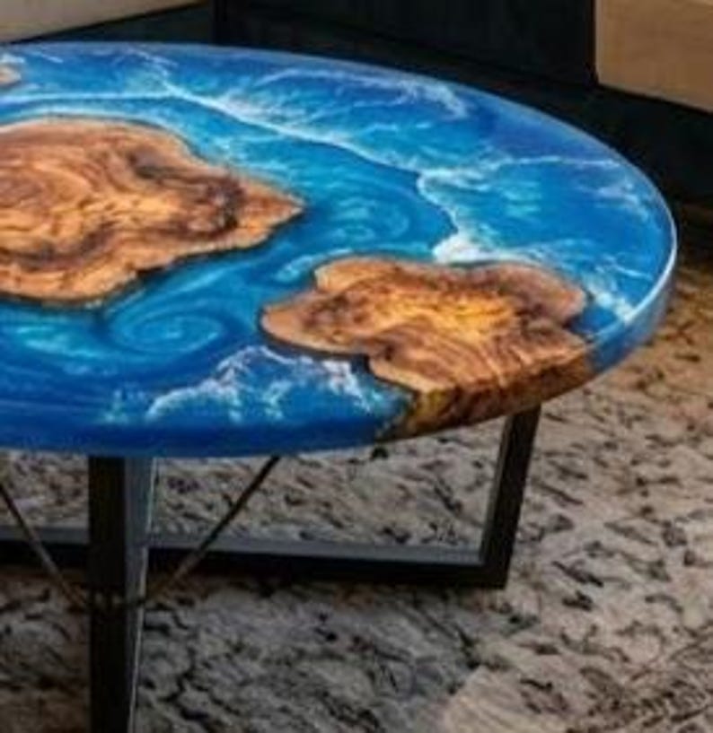 May include: Round coffee table with a blue epoxy resin top, featuring natural wood inlays. The table has a dark metal base. The wood inlays have a natural, organic shape. The table's diameter is approximately 36 inches.