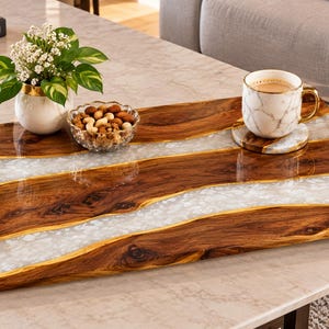 May include: A rectangular serving tray featuring a natural wood grain surface with two wavy, translucent white resin inlays. The tray includes two polished gold-tone metal handles attached to the sides, providing a sleek contrast to the wood.