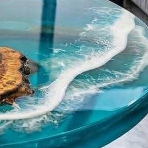 May include: Round epoxy resin table with a blue ocean wave design. The table features a wood burl on the left side, with a clear, glossy finish. The wave design is white and foamy, creating a realistic ocean effect.