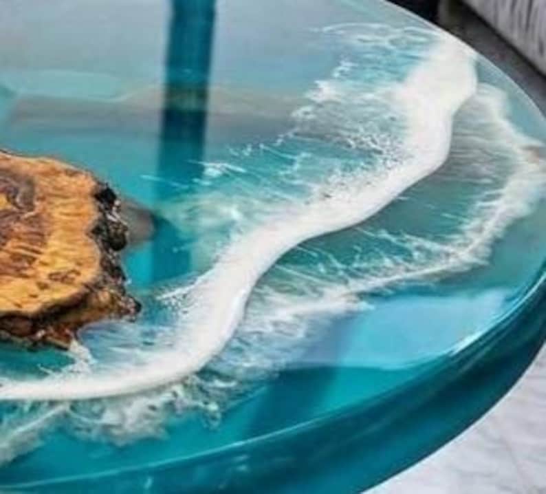 May include: Round epoxy resin table with a blue ocean wave design. The table features a wood burl on the left side, with a clear, glossy finish. The wave design is white and foamy, creating a realistic ocean effect.