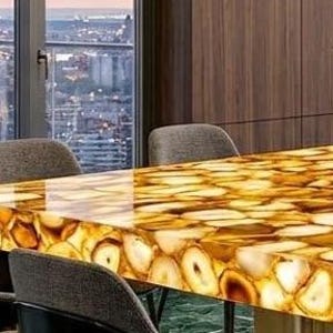May include: A rectangular table with a polished, translucent surface made of golden agate slices. The table is supported by a dark metal frame and surrounded by gray upholstered chairs. The table is approximately 72 inches long.