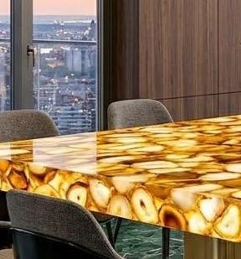 May include: A rectangular table with a polished, translucent surface made of golden agate slices. The table is supported by a dark metal frame and surrounded by gray upholstered chairs. The table is approximately 72 inches long.