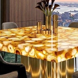 May include: A rectangular table with a golden, agate-like surface. The table has a polished, reflective finish and is supported by gold-colored, cylindrical legs. A vase of dark flowers and gold decorative items sit on the table. The background shows a cityscape.