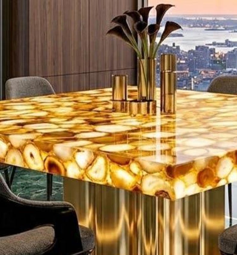 May include: A rectangular table with a golden, agate-like surface. The table has a polished, reflective finish and is supported by gold-colored, cylindrical legs. A vase of dark flowers and gold decorative items sit on the table. The background shows a cityscape.