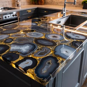 May include: A kitchen countertop with a unique design. The countertop is made of polished agate slices in shades of gray, blue, and white, set in a gold-colored resin. The countertop has a built-in stainless steel sink and is surrounded by dark gray cabinets.