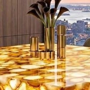 May include: A rectangular dining table with a polished, golden agate stone top. The table is surrounded by gray chairs and set against a backdrop of a cityscape with a bridge. A gold vase with dark flowers and gold candle holders are on the table.
