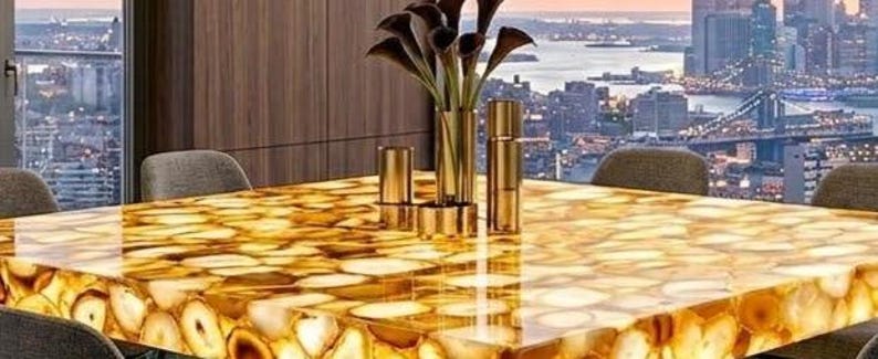 May include: A rectangular dining table with a polished, golden agate stone top. The table is surrounded by gray chairs and set against a backdrop of a cityscape with a bridge. A gold vase with dark flowers and gold candle holders are on the table.