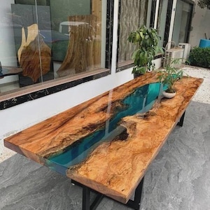 May include: A large, rectangular wooden table with a river-like design. The table features a natural wood grain surface with a vibrant blue epoxy resin river running through the center. The table is supported by black metal legs.