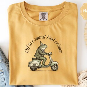 May include: Mustard-coloured t-shirt with the phrase "Off to commit Dad crimes" in a circular arc. The graphic features a frog in a suit riding a scooter. The shirt is folded, with a pair of blue jeans visible on the side.