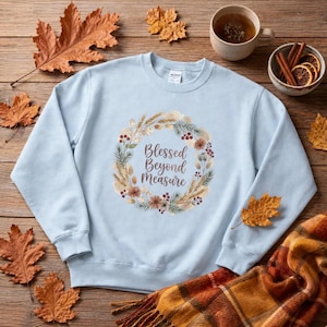 May include: A light blue crewneck sweatshirt featuring a central circular wreath graphic composed of wheat stalks, pine needles, berries, and dried flowers. The text Blessed Beyond Measure is printed in a brown script font inside the wreath.