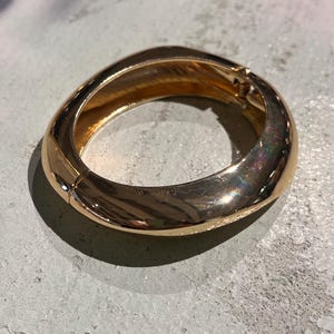 May include: A gold-coloured, oval-shaped hinged bracelet. The bracelet has a smooth, polished exterior and a clasp closure. The design is simple and elegant, with a slight curve to the overall shape. The bracelet is shown on a grey surface.