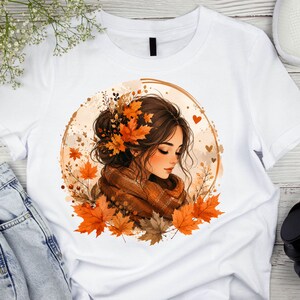 May include: A white t-shirt featuring a circular graphic of a person with brown hair styled in an updo adorned with orange autumn maple leaves. The person wears a brown scarf, surrounded by a warm-toned illustration of falling leaves and hearts.