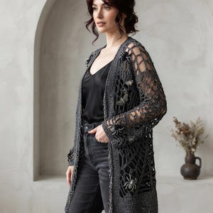 Puede incluir: Un cárdigan de ganchillo abierto de color gris oscuro con un intrincado patrón de telaraña, mangas largas y adornos decorativos de arañas y polillas. La prenda tiene un acabado texturizado. El texto COVEN CROCHET CO. es visible.