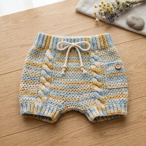 Puede incluir: Pantalones cortos de ganchillo con un patrón de hilo jaspeado en tonos azul claro, amarillo y crema. La prenda incluye una cintura acanalada con cordón funcional, dos paneles verticales de estilo trenzado y un botón de madera decorativo.