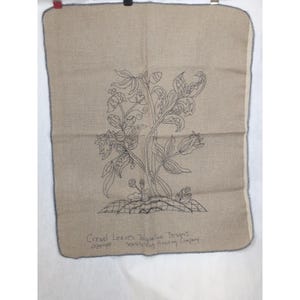 May include: A rectangular piece of tan linen fabric featuring a dark blue line drawing of a leafy plant with flowers. The text Crewel Leaves Jacqueline Designs copyright Seaside Rug Hooking Company is printed in dark blue at the bottom.