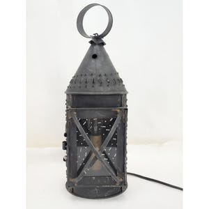 Puede incluir: Un farol de metal negro con una parte superior cónica, un anillo circular para colgar y un panel frontal de vidrio rectangular con un diseño de refuerzo en cruz. El farol tiene una textura de metal perforado y un cable eléctrico negro.