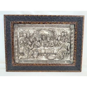 Op de afbeelding: Een rechthoekige zilverkleurige metalen reliëfplaat met een afbeelding van het Laatste Avondmaal met figuren aan een tafel. De tekst "ULTIMA CENA DE JESUS" staat onderaan. De plaat zit in een donkerbruine, getextureerde houten lijst.