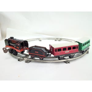 Puede incluir: Un tren de juguete mecánico vintage que incluye una locomotora negra con detalles rojos y llave de cuerda, un vagón de carbón negro, un vagón de pasajeros rojo con techo blanco y un vagón verde, sobre una vía circular de metal.