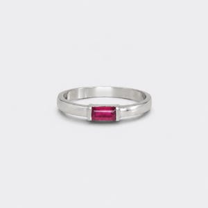 May include: A silver-toned ring featuring a single rectangular baguette-cut deep pink gemstone set horizontally in the center. The band is smooth, polished, and has a consistent width throughout, showcasing a clean and simple metal design.