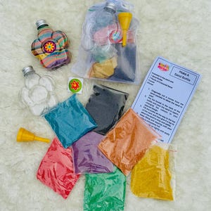 May include: A sand bottle kit with a clear flower-shaped bottle, a yellow funnel, and packets of coloured sand in red, green, orange, yellow, purple, black, and blue. The kit includes instructions and a sticker.