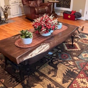 May include: A rectangular coffee table with a dark brown live-edge wooden top and black metal hairpin legs. A red braided table runner sits on the surface, holding a white pot with a green plant and a white bowl filled with red and white flowers.