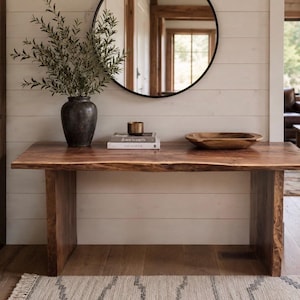 May include: A rectangular live-edge wooden console table with a dark brown finish and two slab legs. On the table sits a dark gray vase with green branches, two stacked books titled Down to Earth, a small metallic cup, and a wooden bowl.