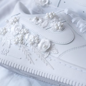 May include: White sneakers with floral and pearl embellishments. The shoes feature delicate, embroidered flowers and pearl accents on the sides and toe. The laces are replaced with white satin ribbons. The word "AIR" is visible on the side of the shoe.