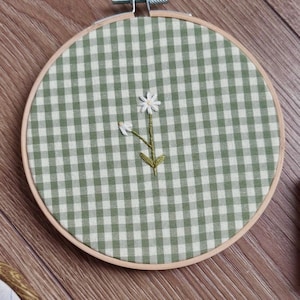 Op de afbeelding: Een ronde houten borduurring met een lichtgroen en wit geruite stof. In het midden van de stof bevindt zich een klein, met de hand geborduurd bloemetje in wit met een geel hartje, een groene steel en twee kleine groene blaadjes.