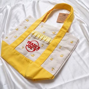 Op de afbeelding: Een crèmekleurige canvas draagtas met gele hengsels en een gele onderkant. De tas is geborduurd met kleine gele citroenmotieven en toont het rode Trader Joe's logo. Een kleine, geplooide gele rand accentueert het voorvak van de tas.