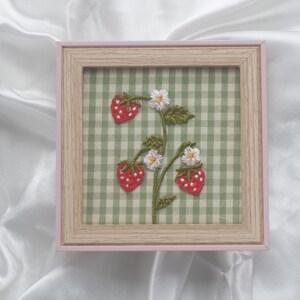 Könnte beinhalten: Ein quadratischer Holzrahmen mit einem Stoff mit grün-weißem Vichy-Muster. Der Stoff ist mit handgestickten roten Erdbeeren mit weißen Samen, grünen Blättern und kleinen weißen Blüten mit gelben Mitten verziert.