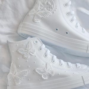 Puede incluir: Par de zapatillas altas blancas adornadas con mariposas. Las zapatillas presentan mariposas bordadas y pequeños detalles de perlas. El diseño clásico de Converse es visible, con cordones blancos y suela de goma.