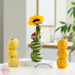 Può includere: Tre vasi in ceramica a forma di frutta: una pila di limoni, angurie e arance. Il vaso di anguria contiene un girasole. I limoni sono gialli, le angurie sono verdi con strisce nere e le arance sono arancioni brillanti.