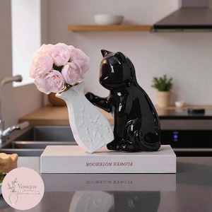 May include: A black ceramic cat ornament supporting a white vase filled with pink flowers. The vase sits on a book with the words "FLOWER NOTEBOOK". The cat has a shiny finish.
