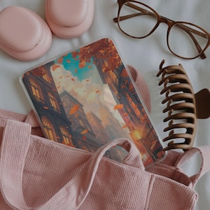May include: A clear tablet case featuring an illustration of a city street during autumn with falling orange leaves, buildings, and a blue sky. The case is placed inside a pink corduroy tote bag next to pink headphones, brown glasses, and a brown hair clip.