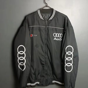May include: Black bomber jacket with white accents and the Audi logo. Features the Audi rings on the sleeve and "S line" text. The jacket has a zip-up front and white striped trim on the collar, cuffs, and hem.