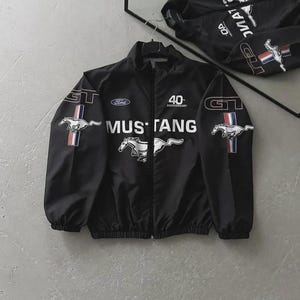 Op de afbeelding: Zwarte Mustang-jas met witte "MUSTANG" tekst en een paardenlogo. Voorzien van "GT" en "Ford" branding, plus een "40th Anniversary" embleem. De jas heeft een volledige ritssluiting en hangt aan een hanger.