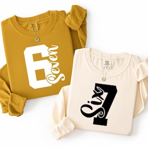 May include: Two long-sleeved sweatshirts. One is mustard yellow with a large white number 6 and the word "Seven" in script. The other is cream-colored with a black number 7 and the word "Six" in script.