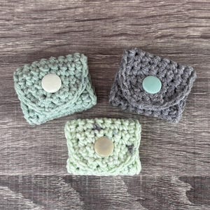 Op de afbeelding: Drie kleine, gehaakte portemonnees in groen- en grijstinten, elk met een knoopsluiting. De portemonnees zijn gerangschikt op een houten oppervlak, wat hun handgemaakte textuur en ontwerp laat zien. De knopen zijn wit en lichtblauw.