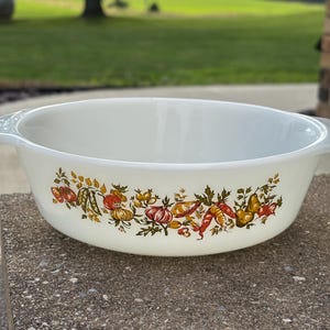 May include: A white round glass casserole dish with integrated side handles. The exterior features a colorful harvest-themed pattern of various vegetables including tomatoes, carrots, onions, and peppers in shades of red, orange, yellow, and green.