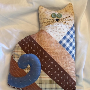 May include: A handmade patchwork cat-shaped pillow. The cat features a cream-colored head with button eyes and beaded whiskers. The body is made of various patterned fabrics in brown, blue, and cream, with a blue swirl tail.