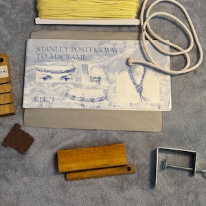 May include: A macrame kit with yellow cord, a white rope, wooden tools, and a metal clamp. The kit includes instructions titled "Stanley Postek's Way to Macrame" and a small brown felt piece. The kit is on a gray surface.